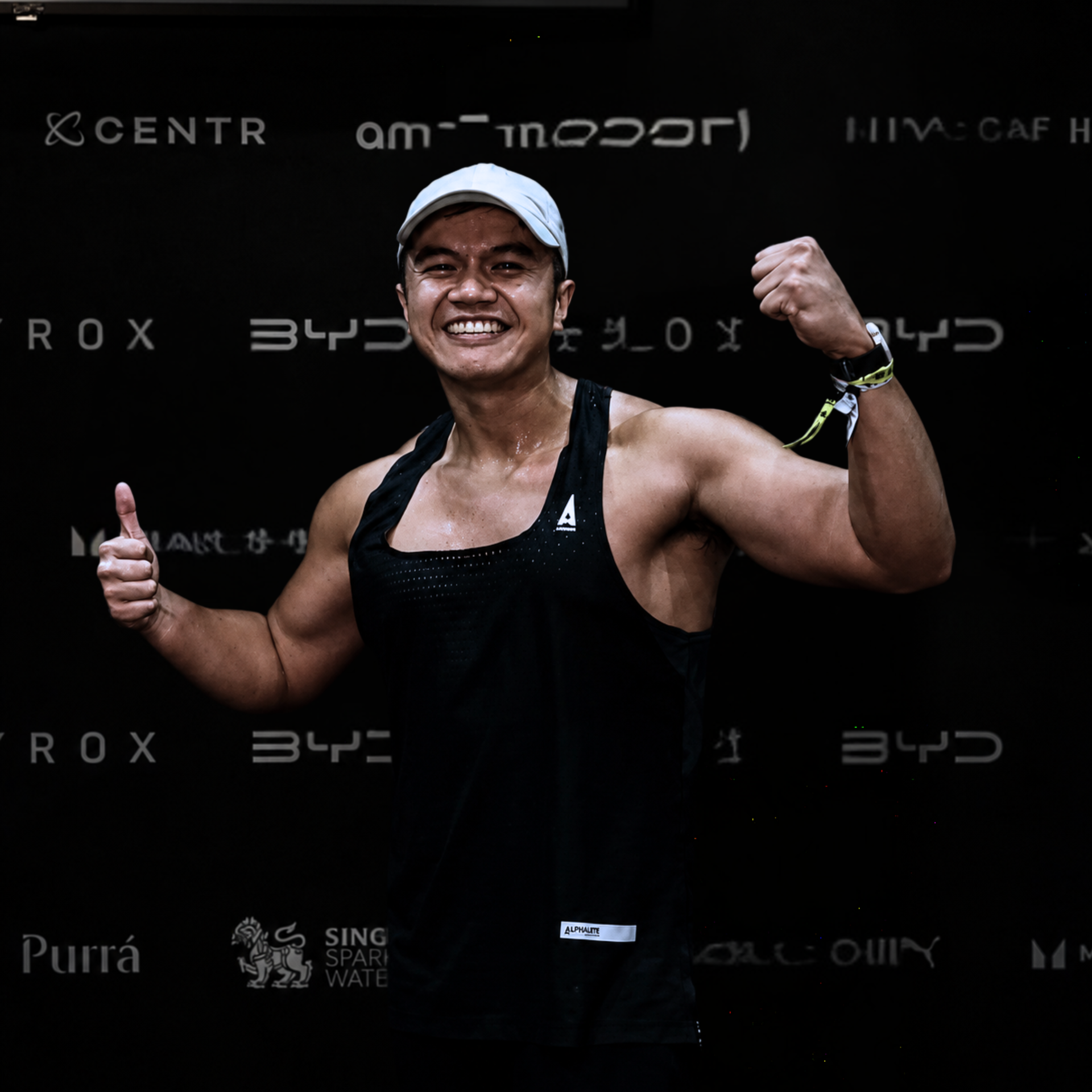 Smiling athlete in black tank top flexing and giving thumbs up against a dark sponsor backdrop