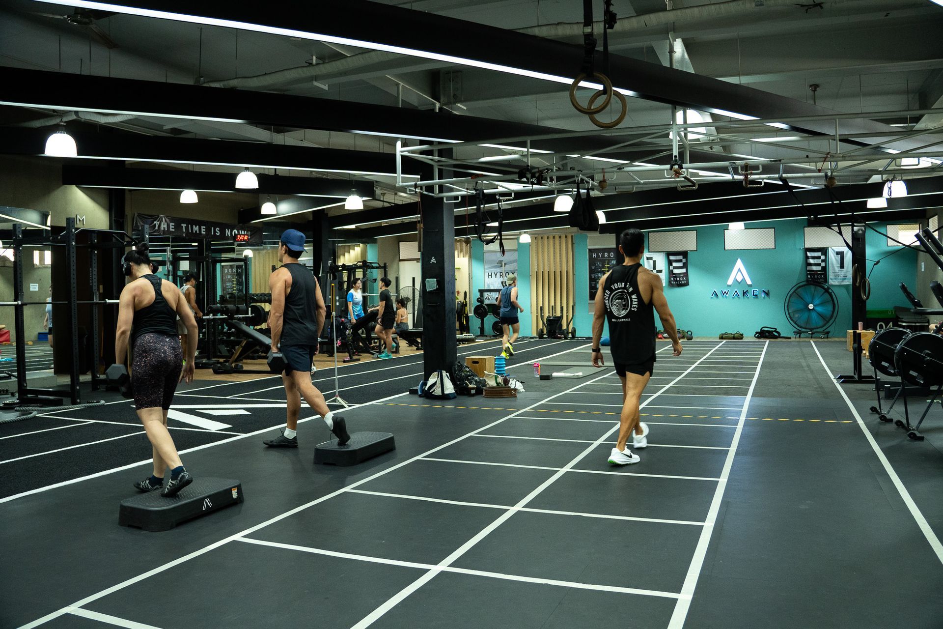 People in matching yellow shirts exercise in a modern gym featuring a green turf track with white floor markings.