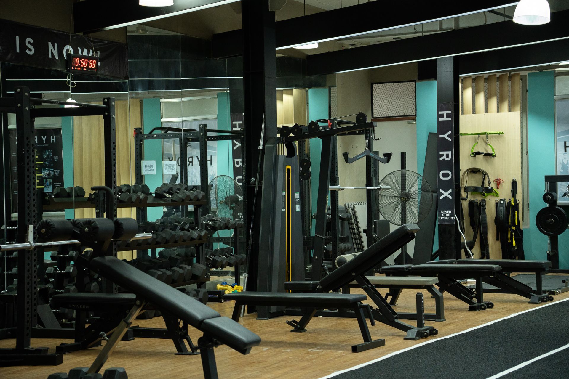 Gym interior with weight benches, barbells, and mirrors, dimly lit with black and teal equipment.