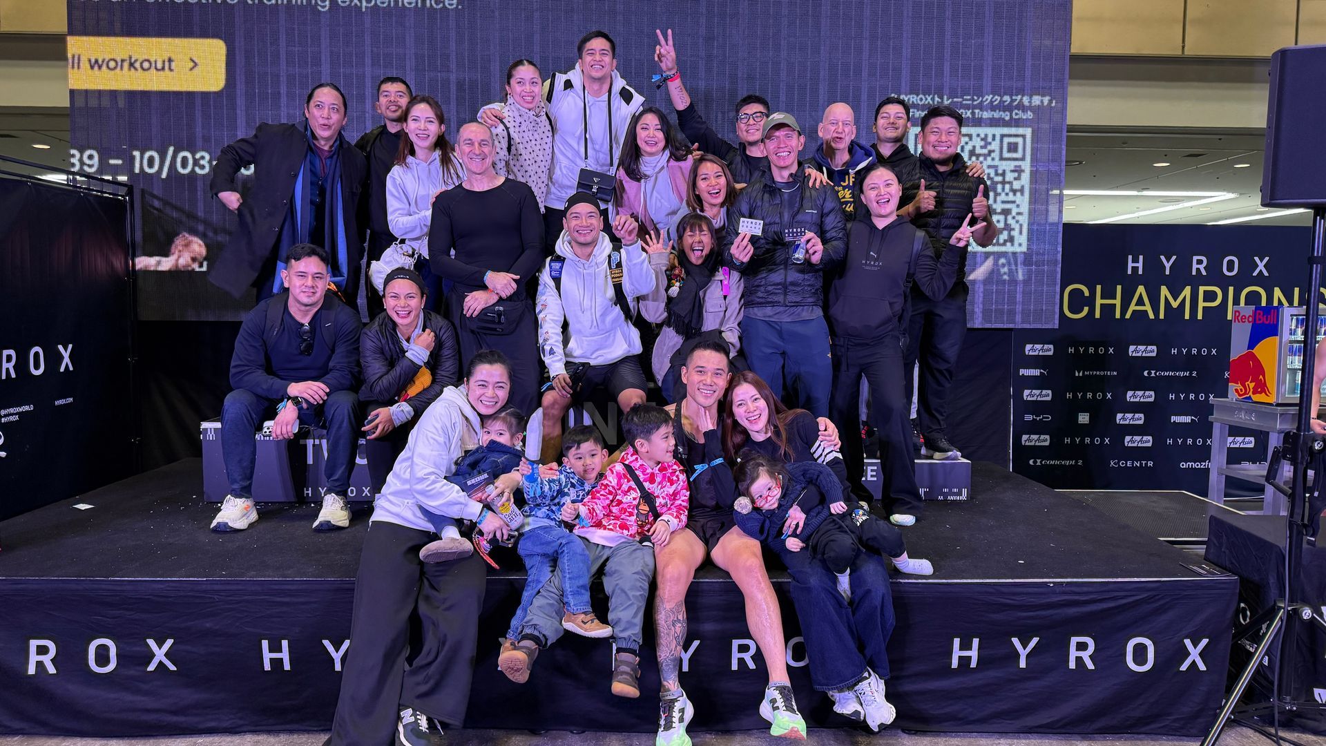 Group posing on a HYROX podium with a trophy and medals in an indoor event hall