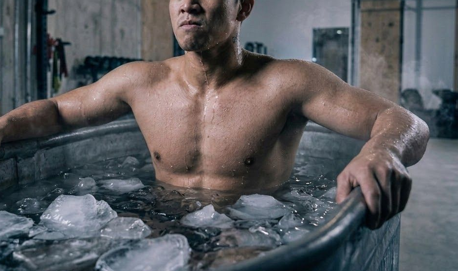 A person with a neutral expression sits in a metal tub filled with ice water in an industrial gym setting.