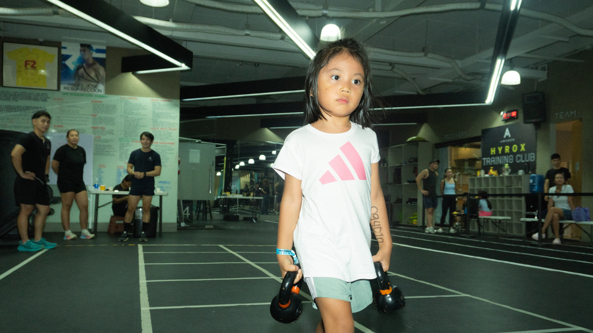 Child holding dumbbells in a gym, standing on a workout floor under bright lights.