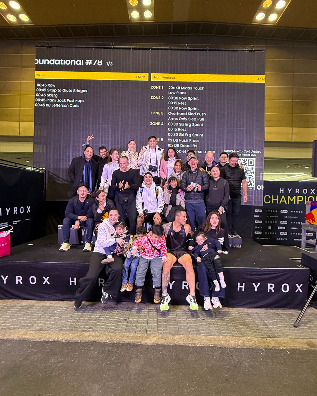 Group of people posing on a HYROX podium with a black-and-yellow event backdrop at night.