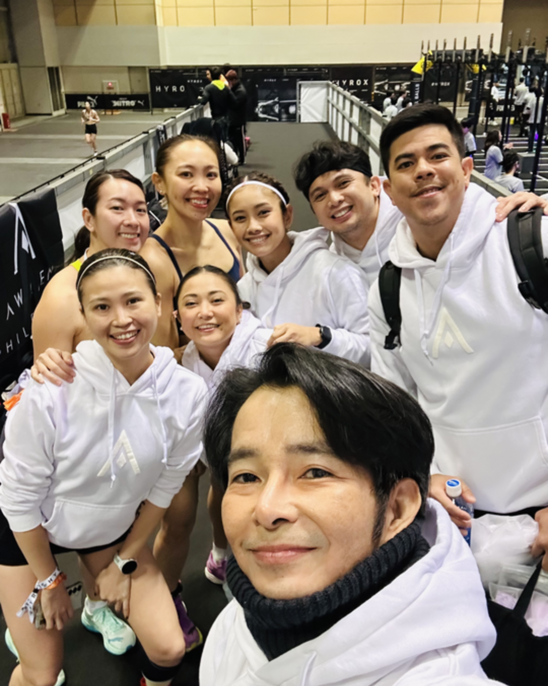 Group of six smiling people in white hoodies taking a selfie indoors, likely at a gym or arena.