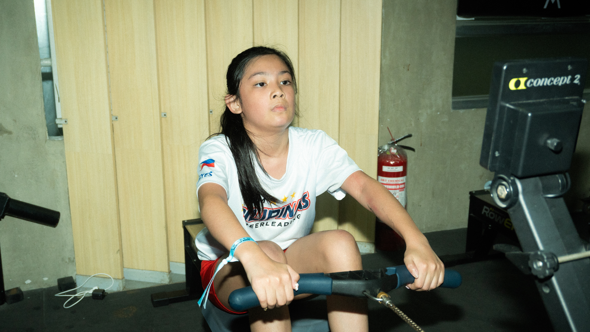 Woman using a rowing machine in a gym, focused and leaning forward, with wood-paneled walls behind her