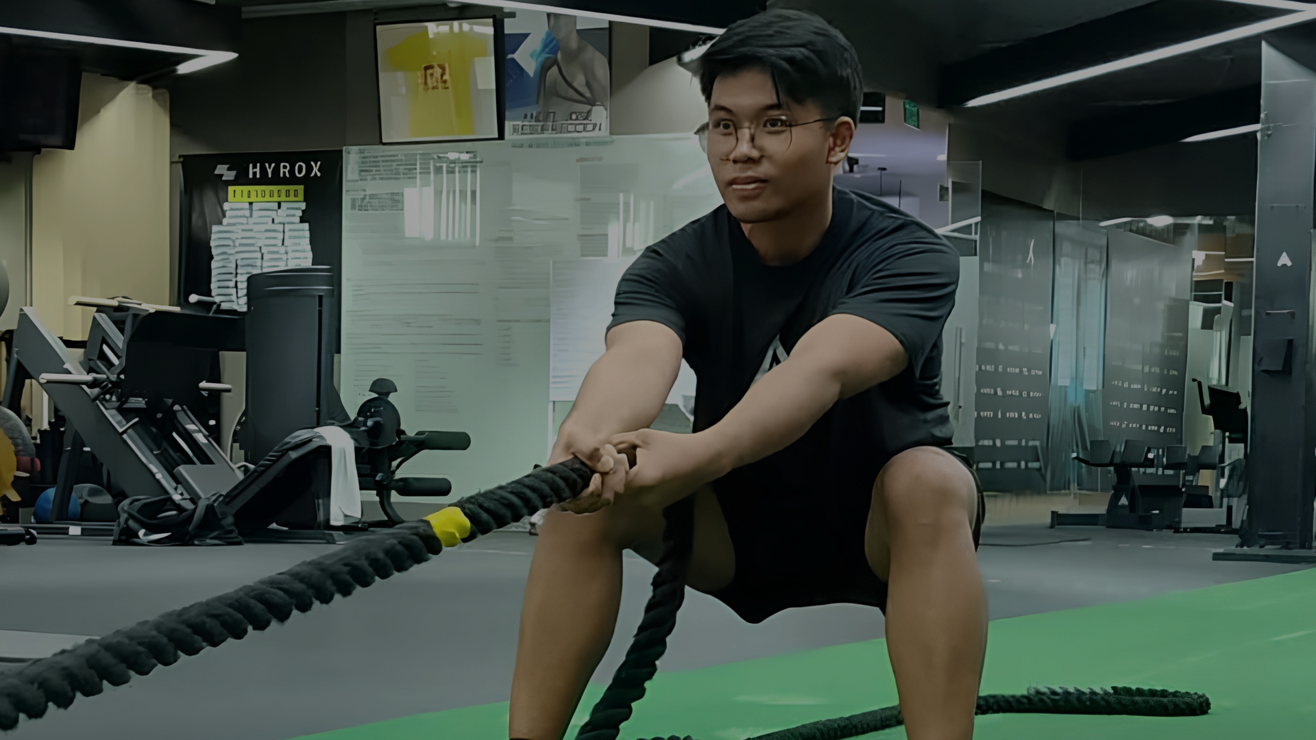Person exercising with battle ropes in a gym, seated on a green mat.