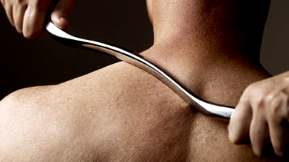 A practitioner uses a metal tool to perform Graston Technique therapy on a person's upper back.