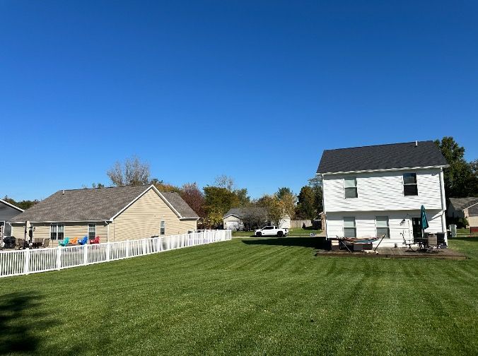 Backyards of two houses, with green lawns, white fences, and a bright blue sky.