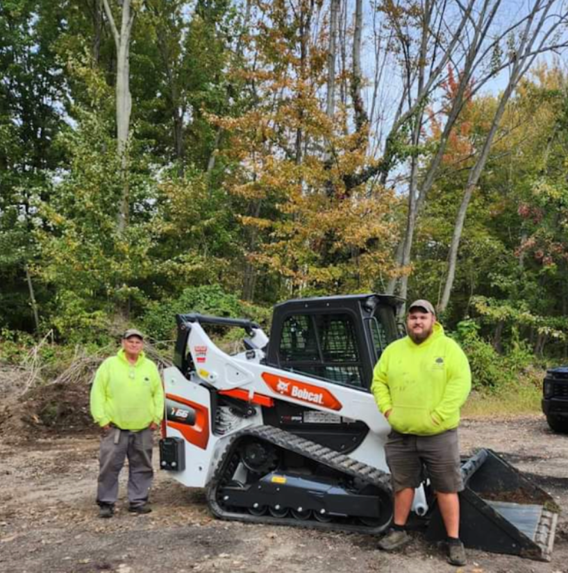 Two men in neon shirts stand near a white Bobcat track loader in a wooded area.
