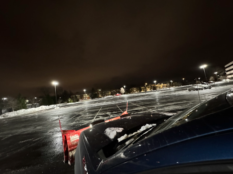 A dark, snowy parking lot at night; a vehicle with a plow blade is in the foreground.