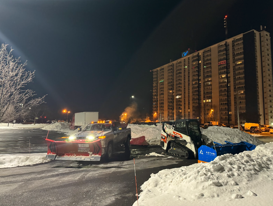 Snowplow clearing a parking lot next to a high-rise building at night.