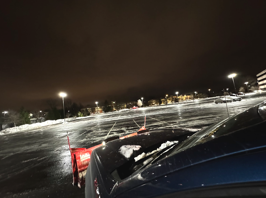 Snowplow attached to a dark blue truck in a snowy parking lot at night. Streetlights illuminate the scene.