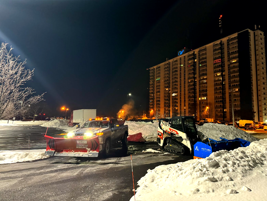 Snowplow and skid steer clearing snow in a parking lot at night, near a tall apartment building.