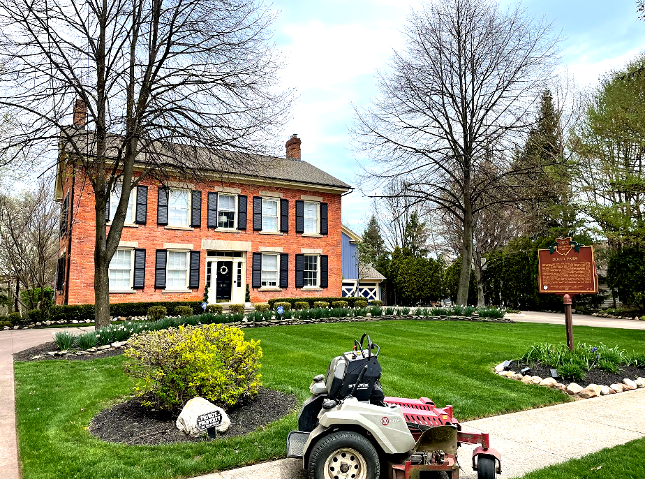 A red brick house with black shutters, a green lawn, and a lawnmower. Historical marker visible.