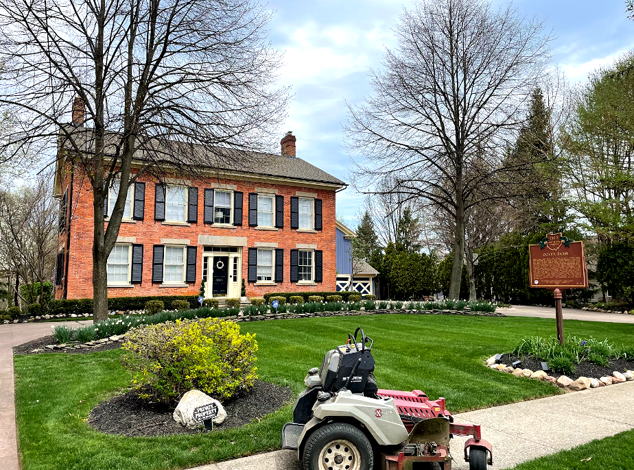 Red brick house with black shutters, lush green lawn, lawnmower, and a historical marker.