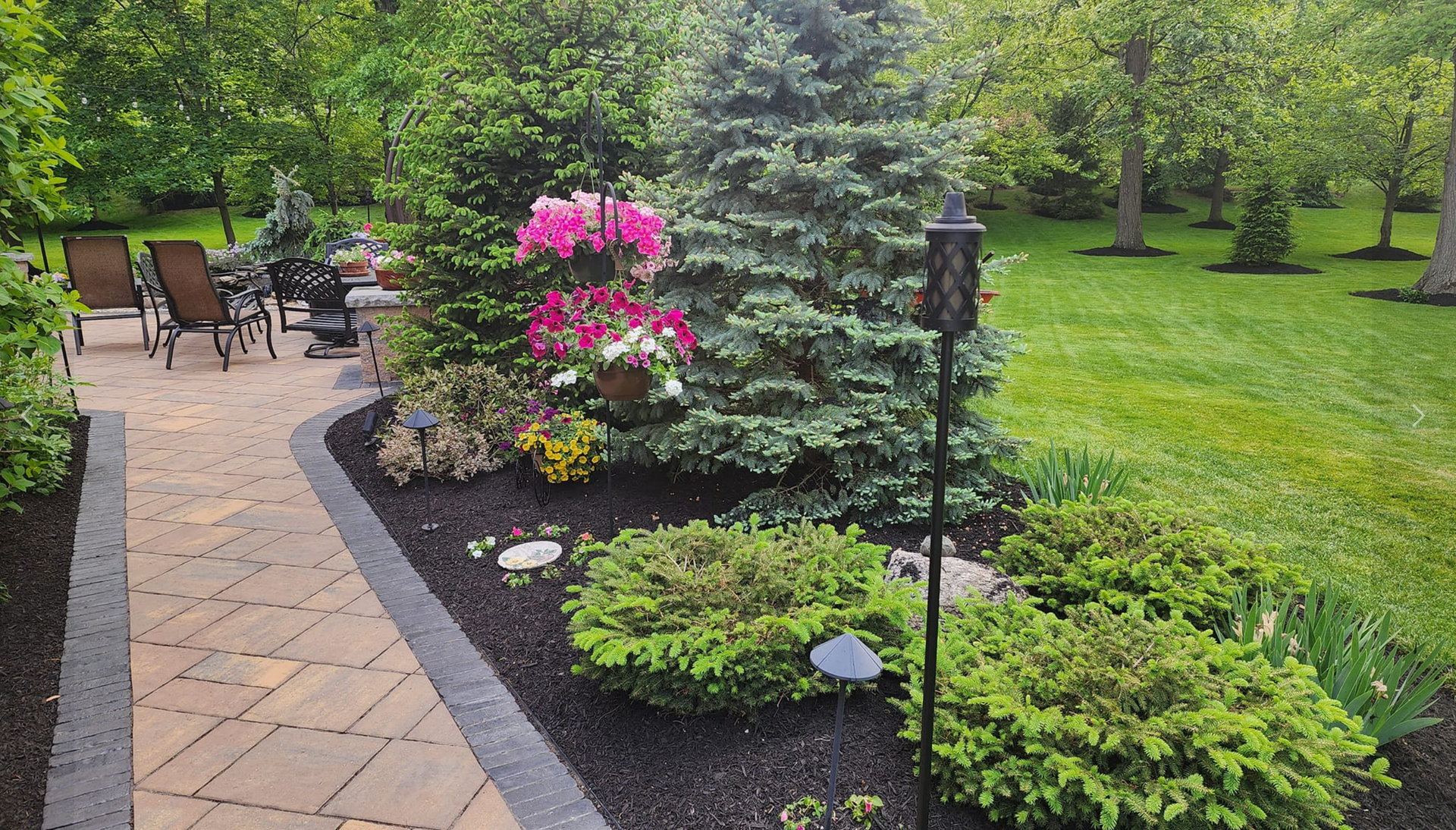 Brick patio with garden bed filled with green bushes and colorful flowers. Trees in the background.