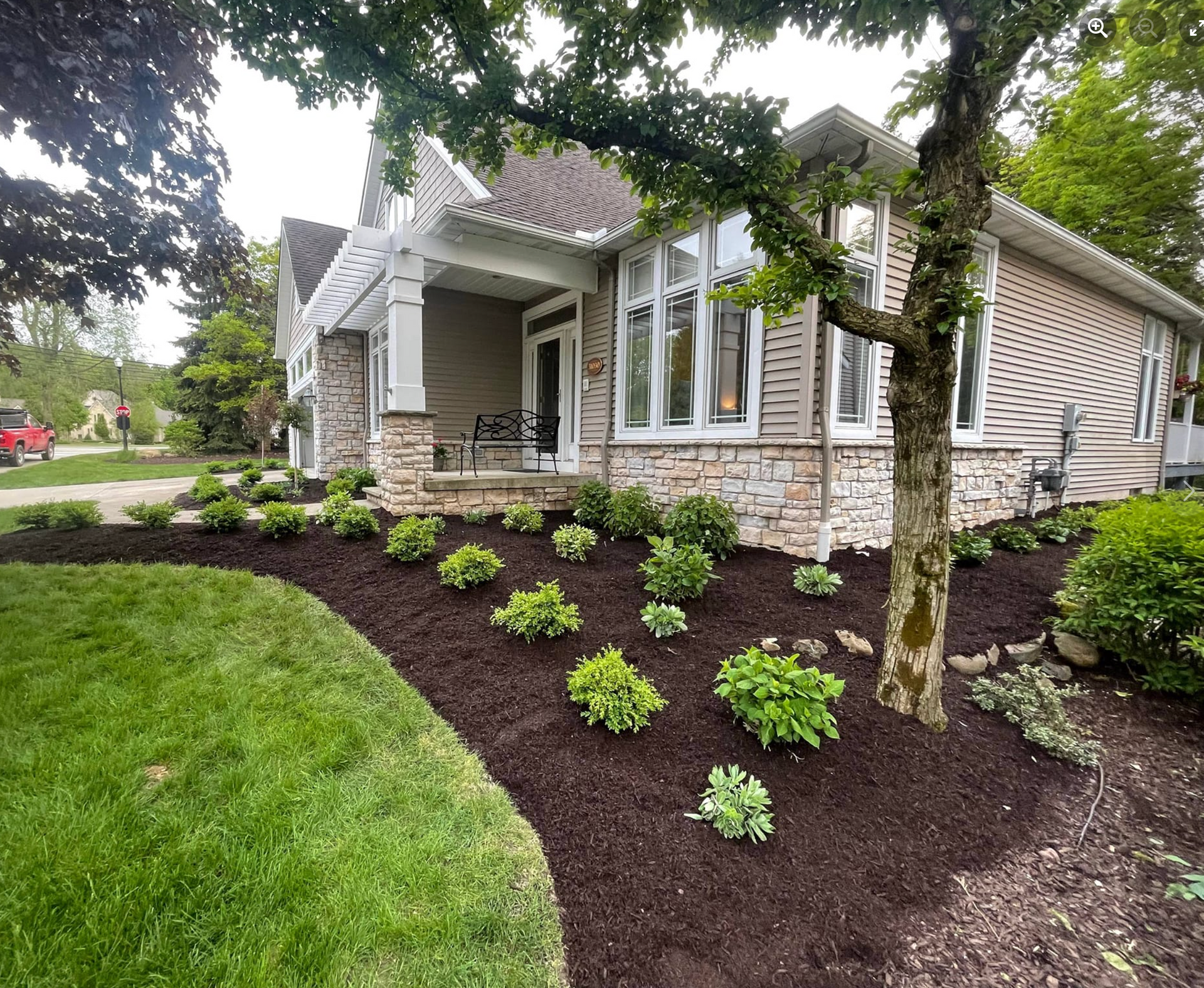 A house with newly landscaped flower beds, mulched in brown, with green plants, and a tree.