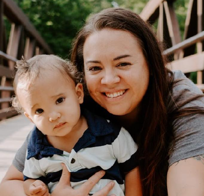 Woman smiles, holding a young child. They sit on a wooden bridge, outdoors.