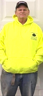 Man in a yellow hoodie, baseball cap, and jeans, standing in front of a white wall.