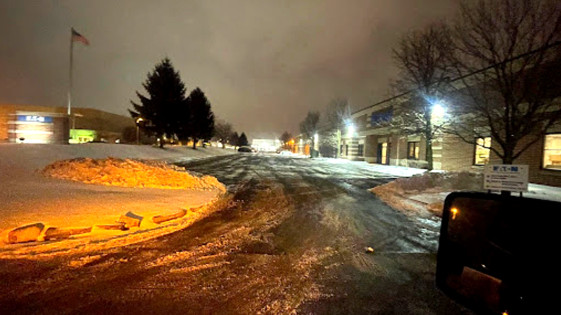 Snowy industrial street at night, with buildings, trees, and a flag. Orange streetlights reflect off the snow.