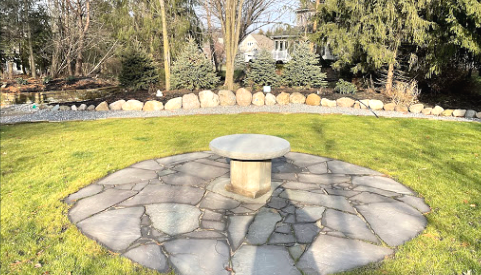 Stone patio with a mushroom-shaped centerpiece, surrounded by grass and a rock border, trees in background.