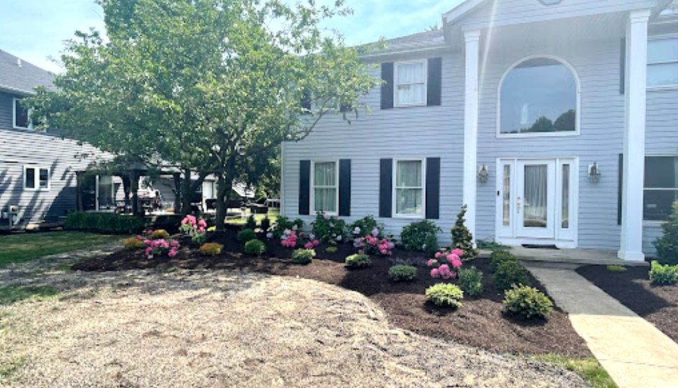 Two-story house with light blue siding, black shutters, and a well-landscaped flower bed with pink blooms.