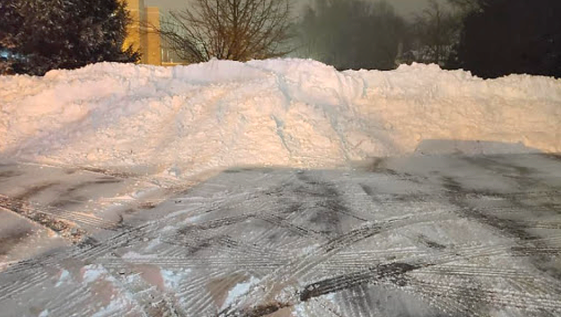 Large pile of snow in a parking lot, illuminated by streetlights, with tire tracks in the snow.