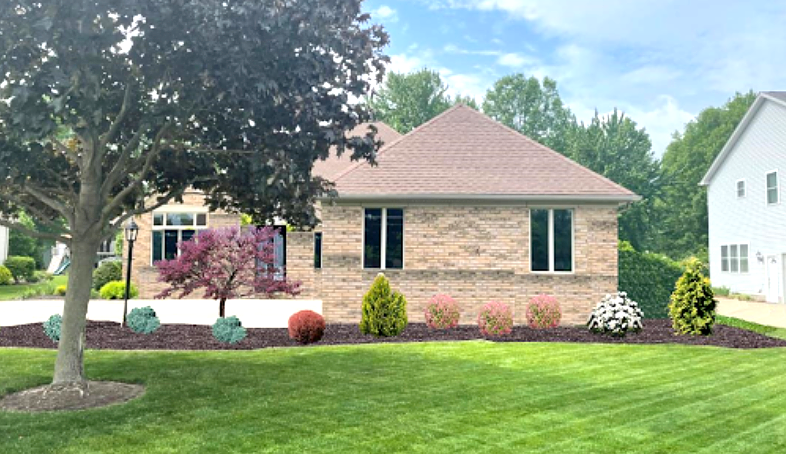 Brick house with brown roof, green lawn, and landscaping featuring colorful shrubs and trees.