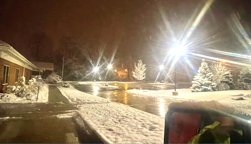 Snowy night scene with a building on the left and bright lights reflecting on wet ground. Snow covers trees and walkways.