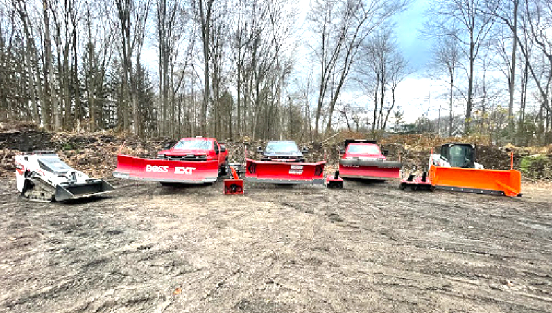 Snowplows lined up outdoors on a muddy surface, with a forested background.