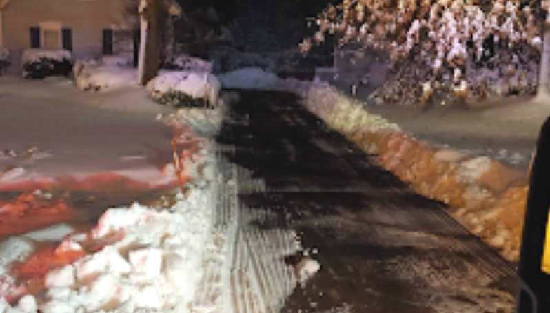 Snow-covered residential street at night, cleared path through the middle, snow piled on both sides, house in background.