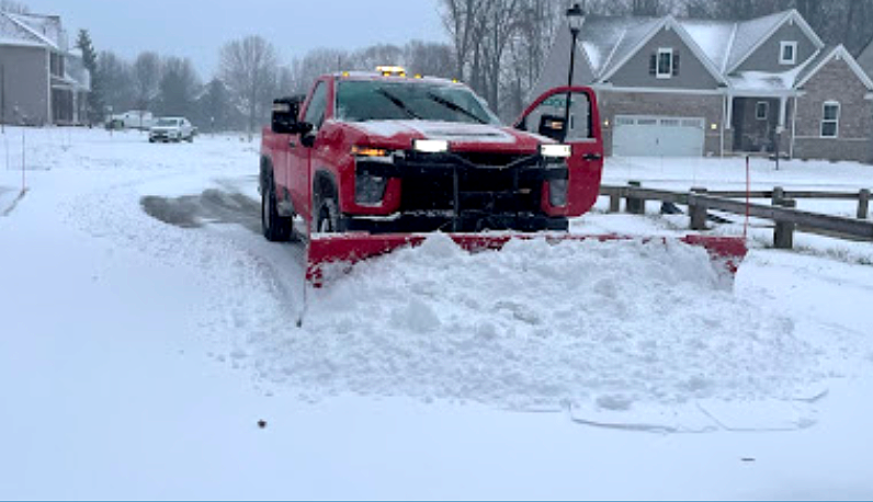 Red pickup truck with snowplow clearing a snow-covered suburban street; door open.