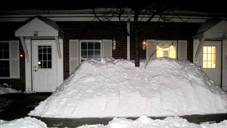 Houses with snow piled high in front of them, covering doorways and windows. Dark night setting.