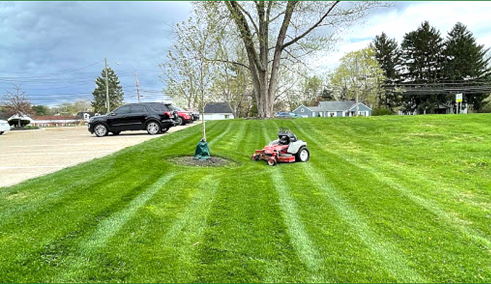 A riding lawnmower cuts grass on a sloped lawn, creating striped patterns. A black SUV is parked nearby.