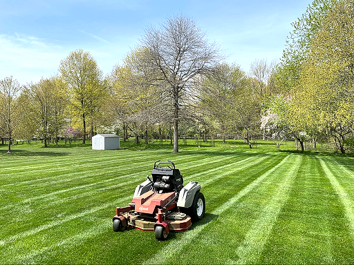 Lawn mower cutting a bright green lawn with trees in the background under a blue sky.