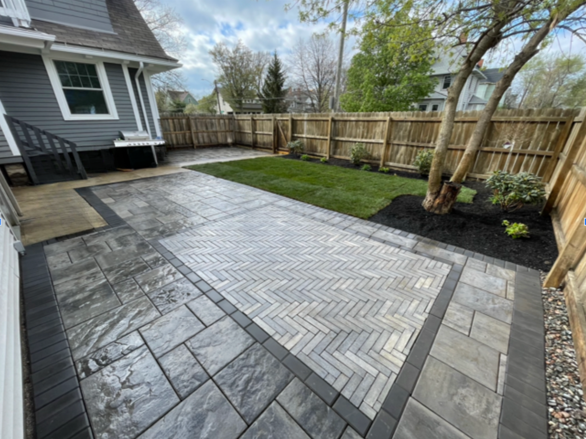 Backyard patio with herringbone pattern, surrounded by dark pavers, grass, and wooden fence.