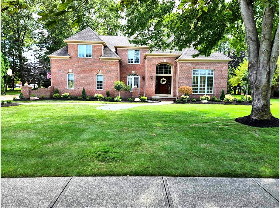Red brick two-story house with green lawn, trees, and sidewalk in front.