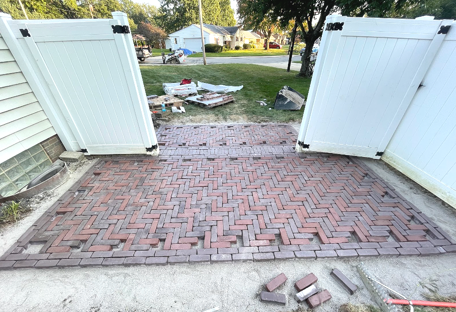 Brick patio between two white fences. Yard work materials nearby.