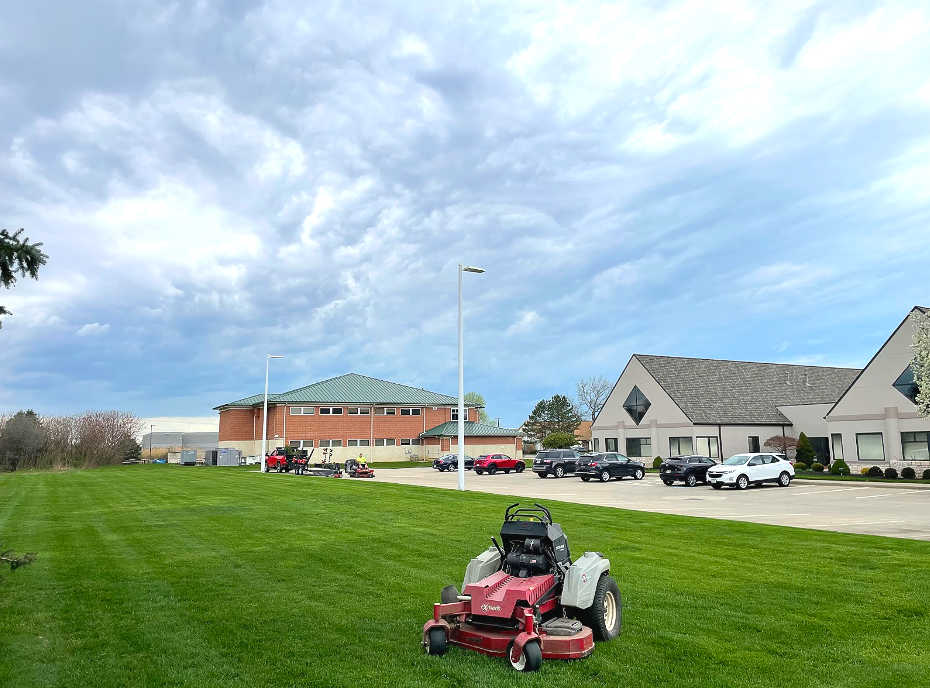 A red and white riding lawnmower on a freshly cut green lawn near a building with cars parked in the lot.