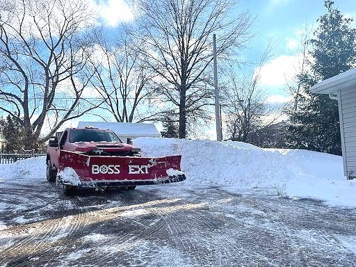 Red pickup truck with a snow plow clears snow, in front of a house, sunny winter day.
