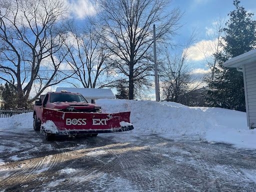 Red pickup truck with a snowplow pushing a large snow pile on a driveway, cloudy sky.