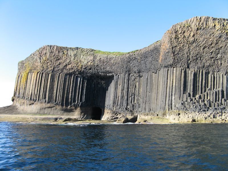 Coastal cliffs of black basalt columns with a sea cave opening, under a blue sky.
