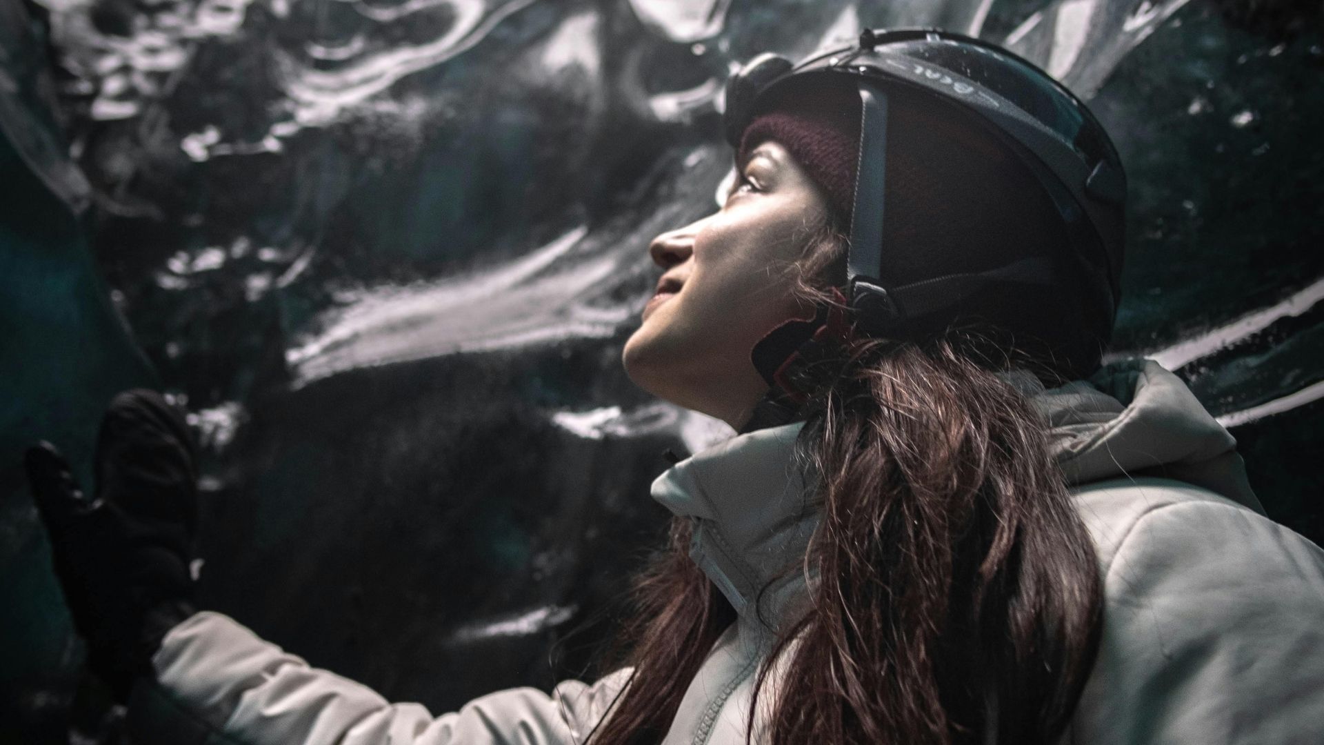 Woman in helmet and jacket looking up at a glacier, holding the ice.
