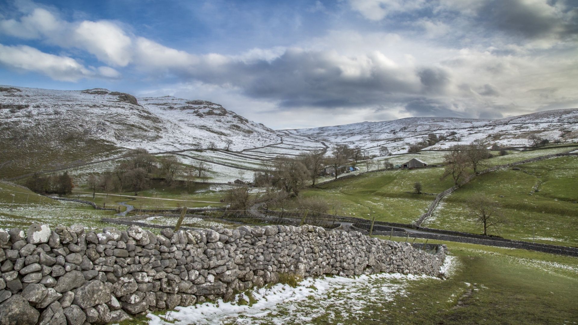 Snow-dusted hills and green fields separated by stone walls under a cloudy sky.
