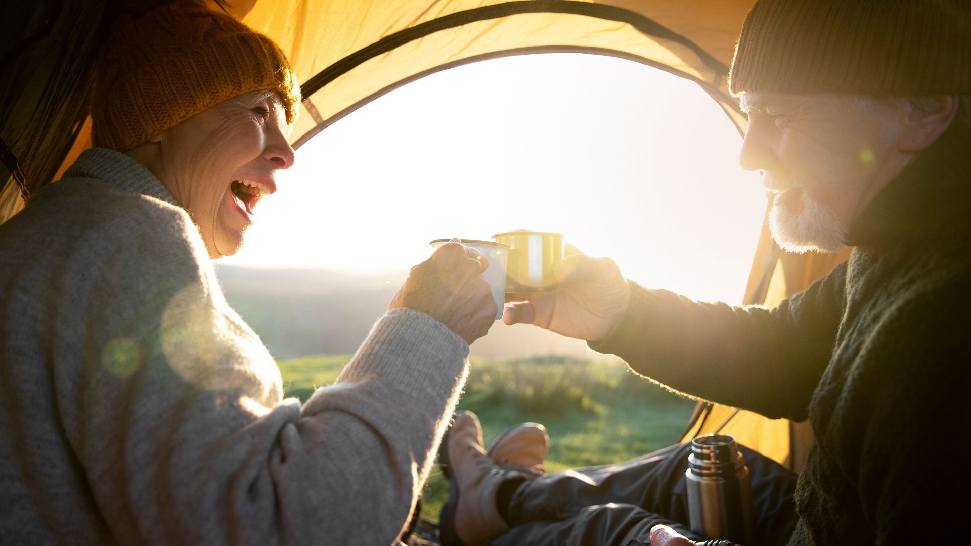 Couple camping, toasting mugs in tent, illuminated by sunlight.