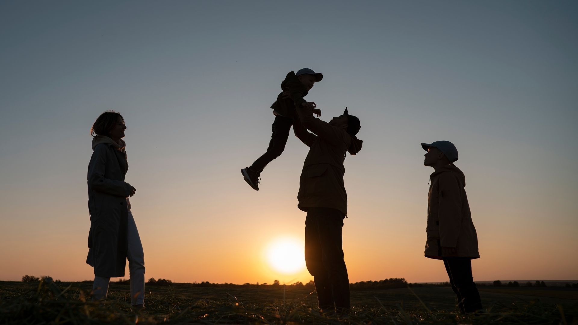 Family silhouetted against a sunset; father throws child in the air, while the mother and another child watch.