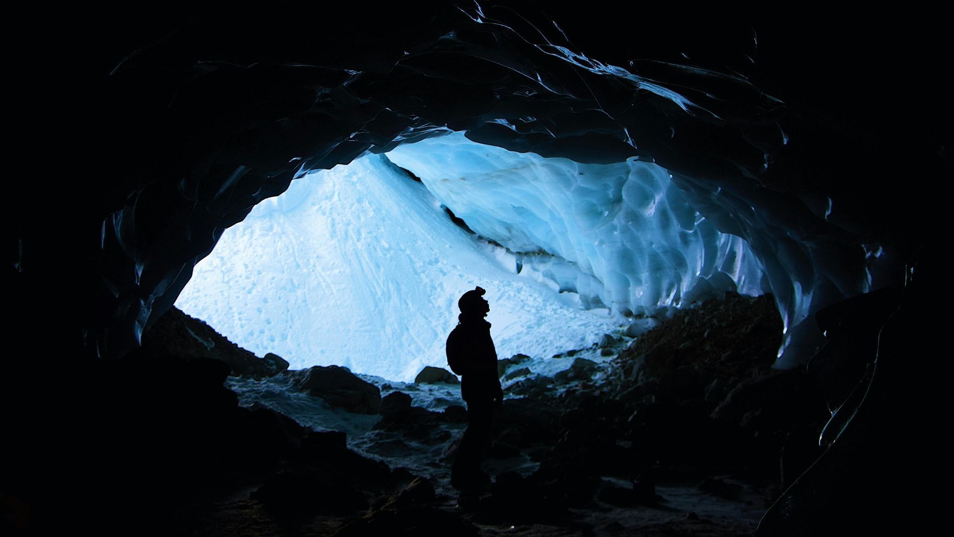 Person silhouetted in a dark cave entrance, looking out at a blue ice formation.