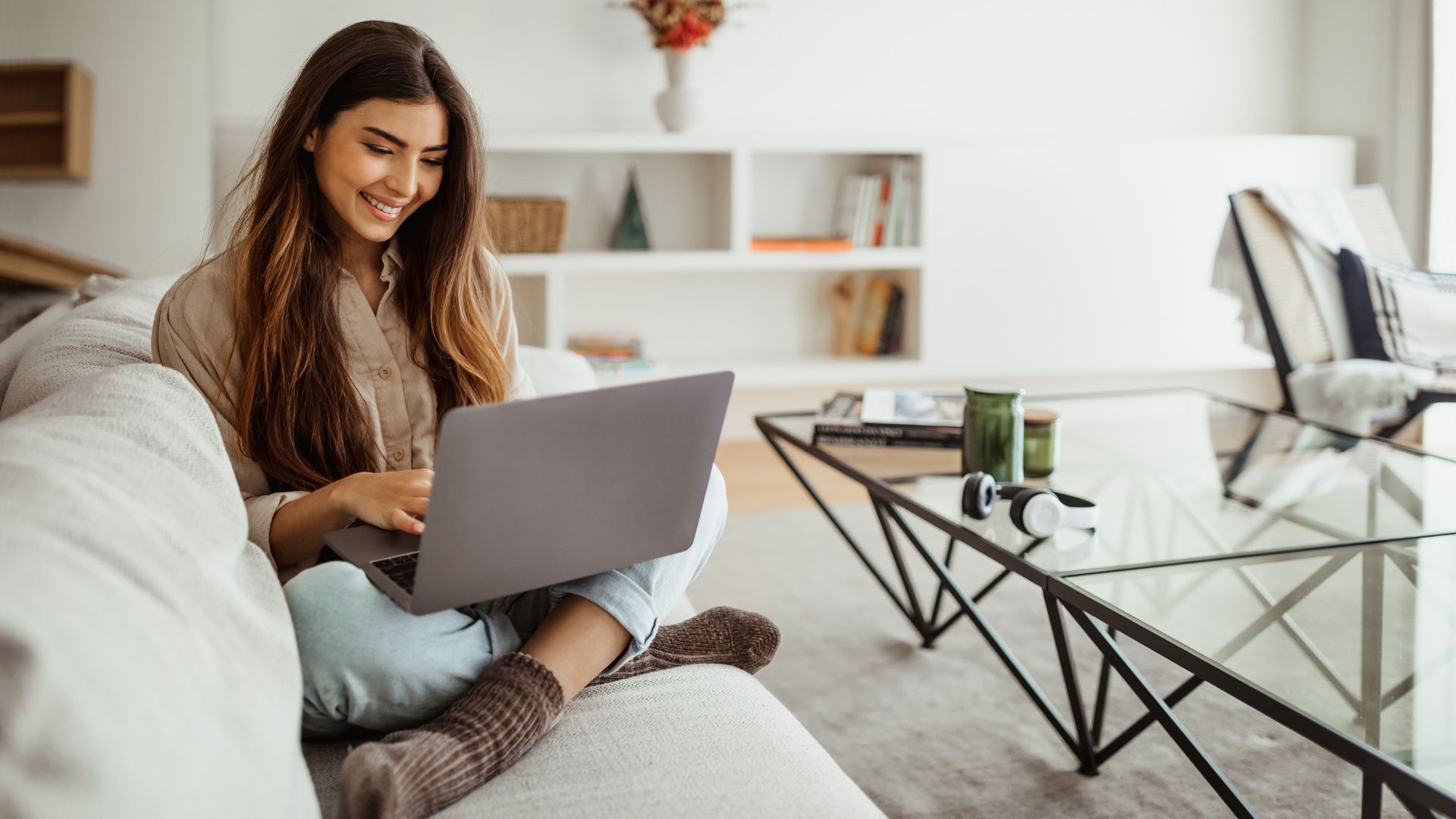 Woman smiling, using a laptop while sitting on a couch in a bright living room.