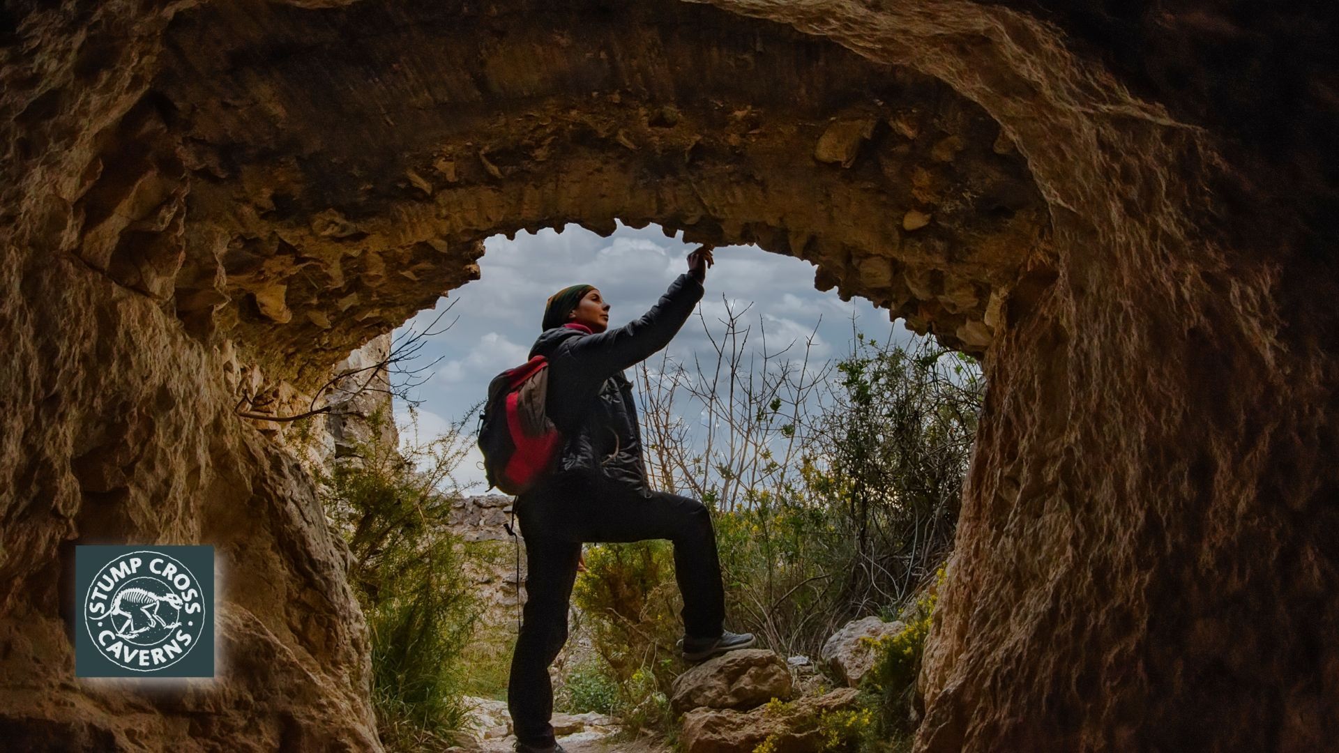 Person touching the top of a cave opening, wearing a backpack, in a natural outdoor setting with a cloudy sky.