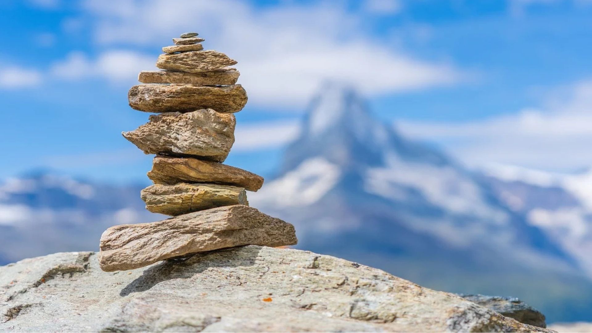 Stone cairn on rock with a blurred mountain and blue sky background.